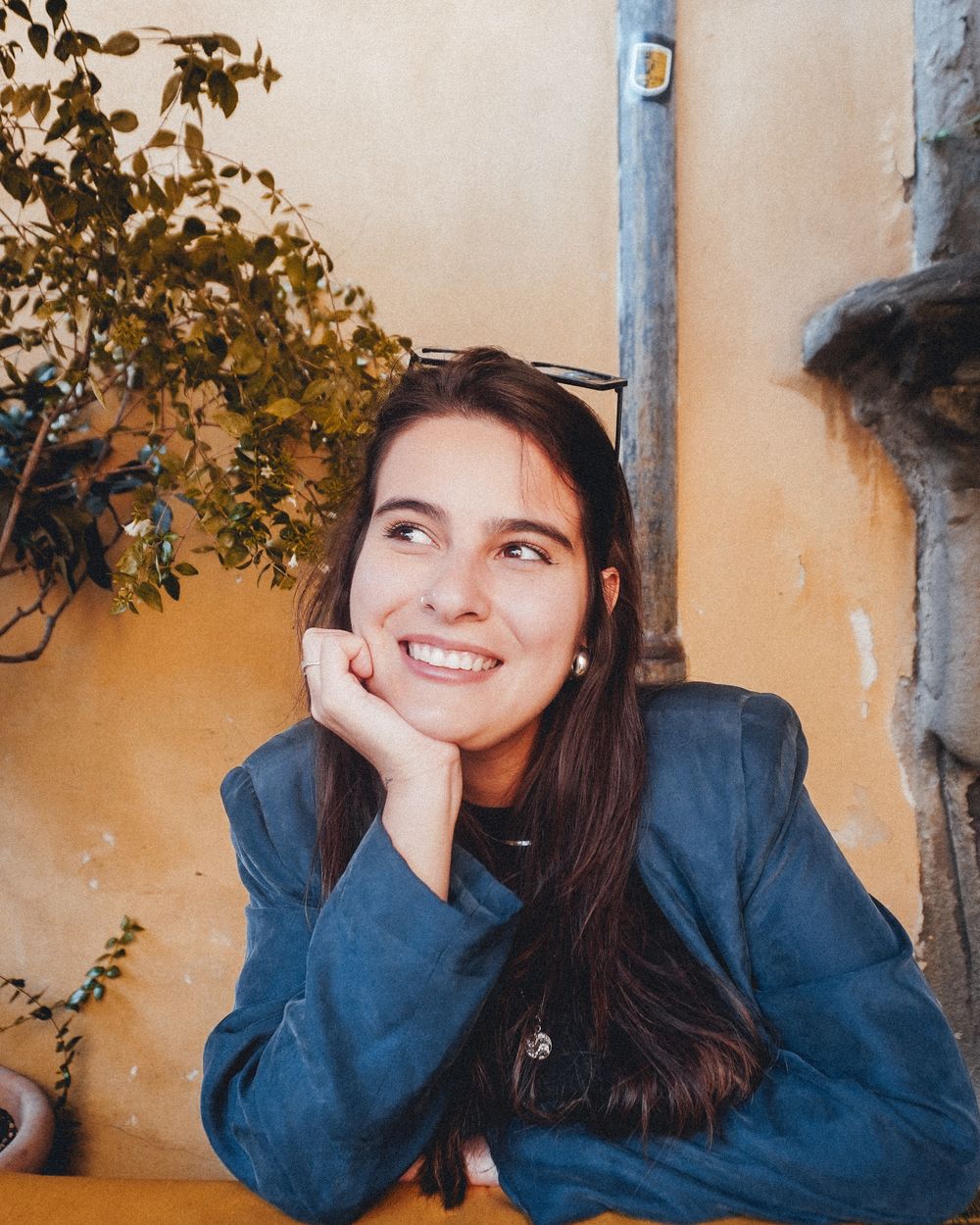 Anna Tamaio sorrindo, com a mão apoiada no rosto, diante de uma parede ocre